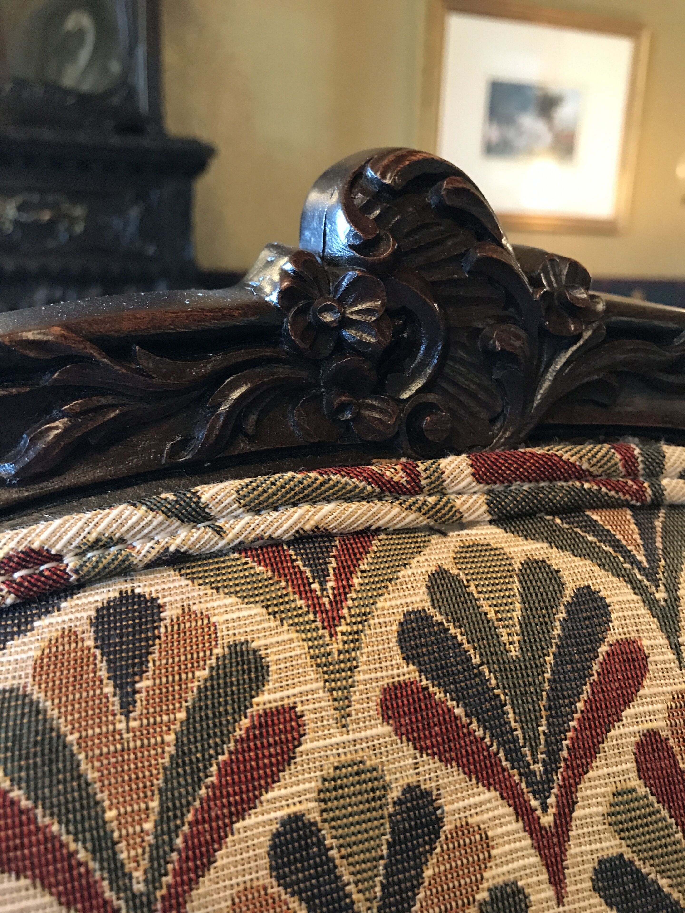 A close-up of a dark wooden furniture piece with intricate carvings, a patterned fabric with a leaf design, and a framed picture in the background.