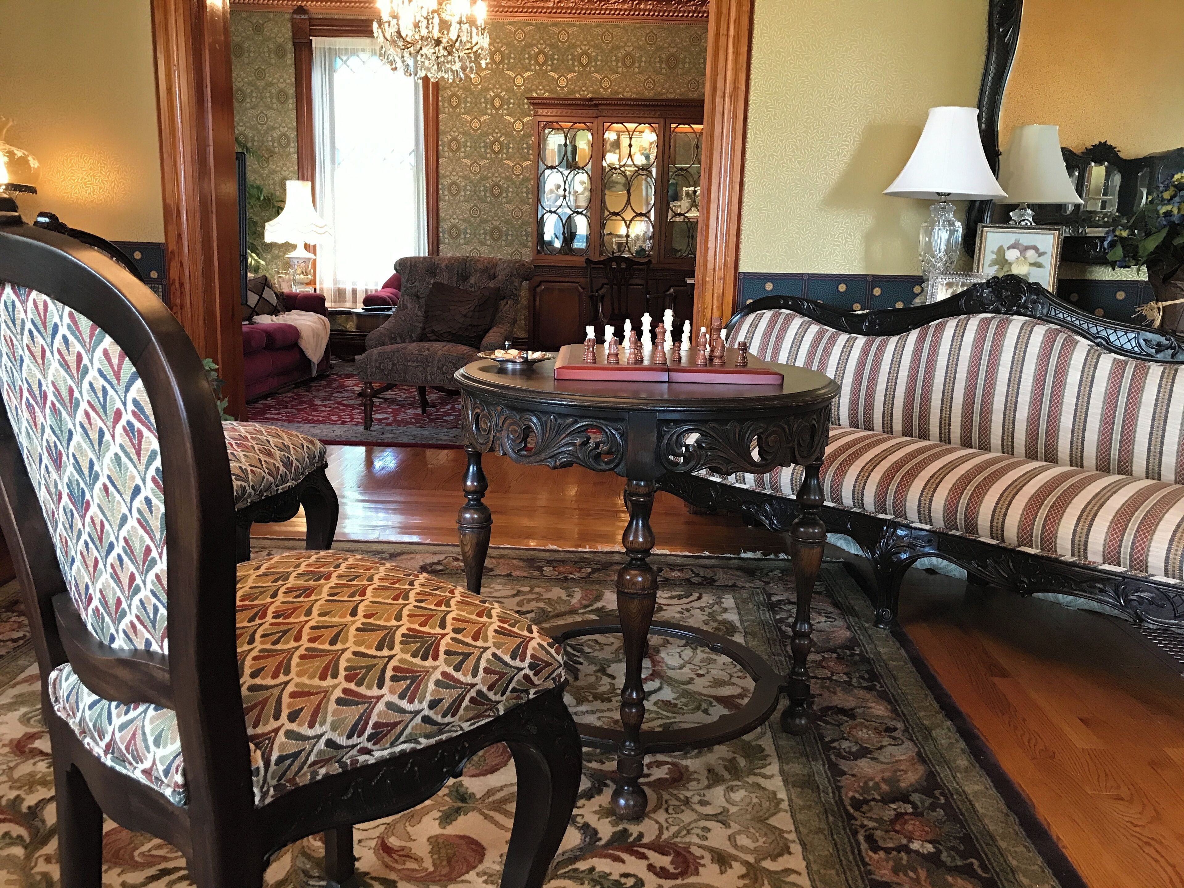 Un salón tradicional con un sillón estampado, un sofá a rayas, una mesa redonda con piezas de ajedrez y una araña.