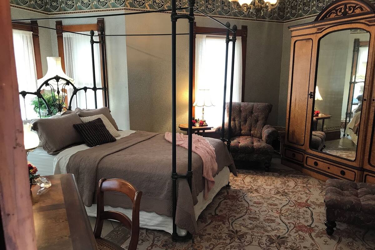 Ein Schlafzimmer mit einem Himmelbett, einem Holzstuhl, einem Schreibtisch und einem großen Spiegel.