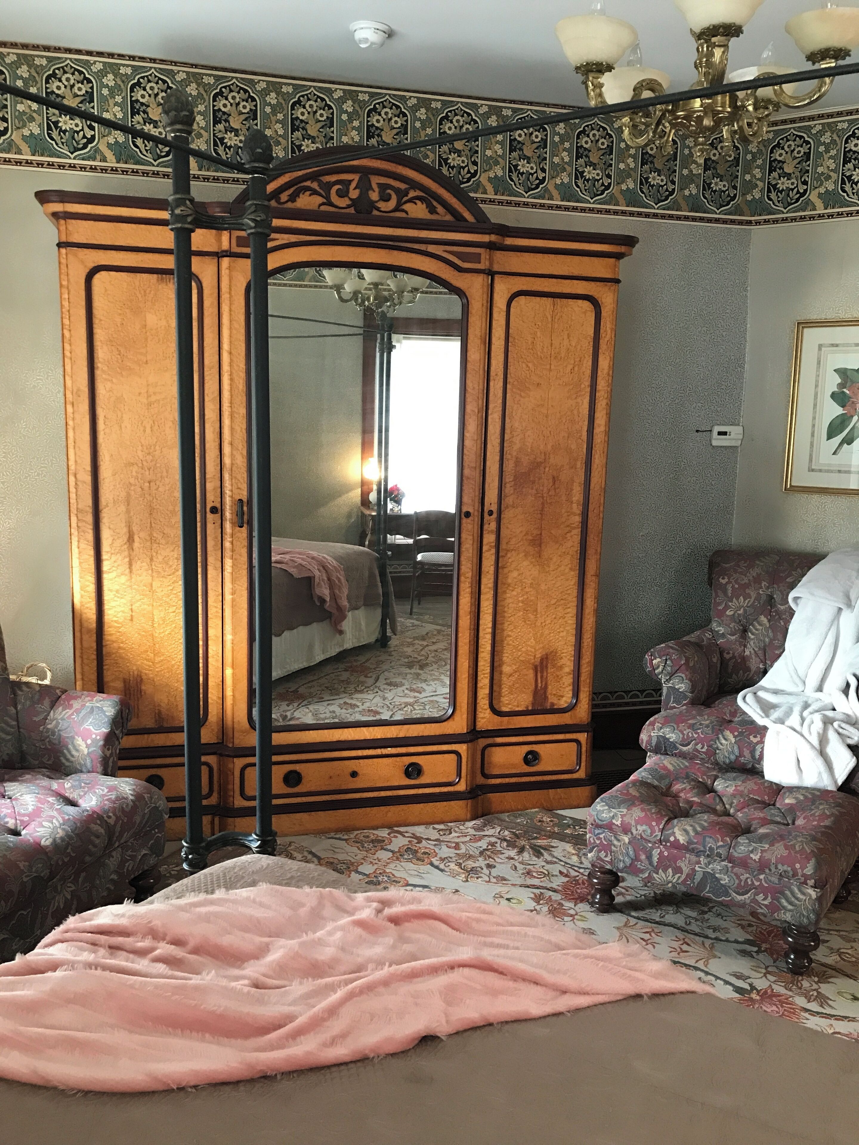 A bedroom with a large wardrobe, floral-patterned armchairs, a bed with a pink blanket, and a chandelier.