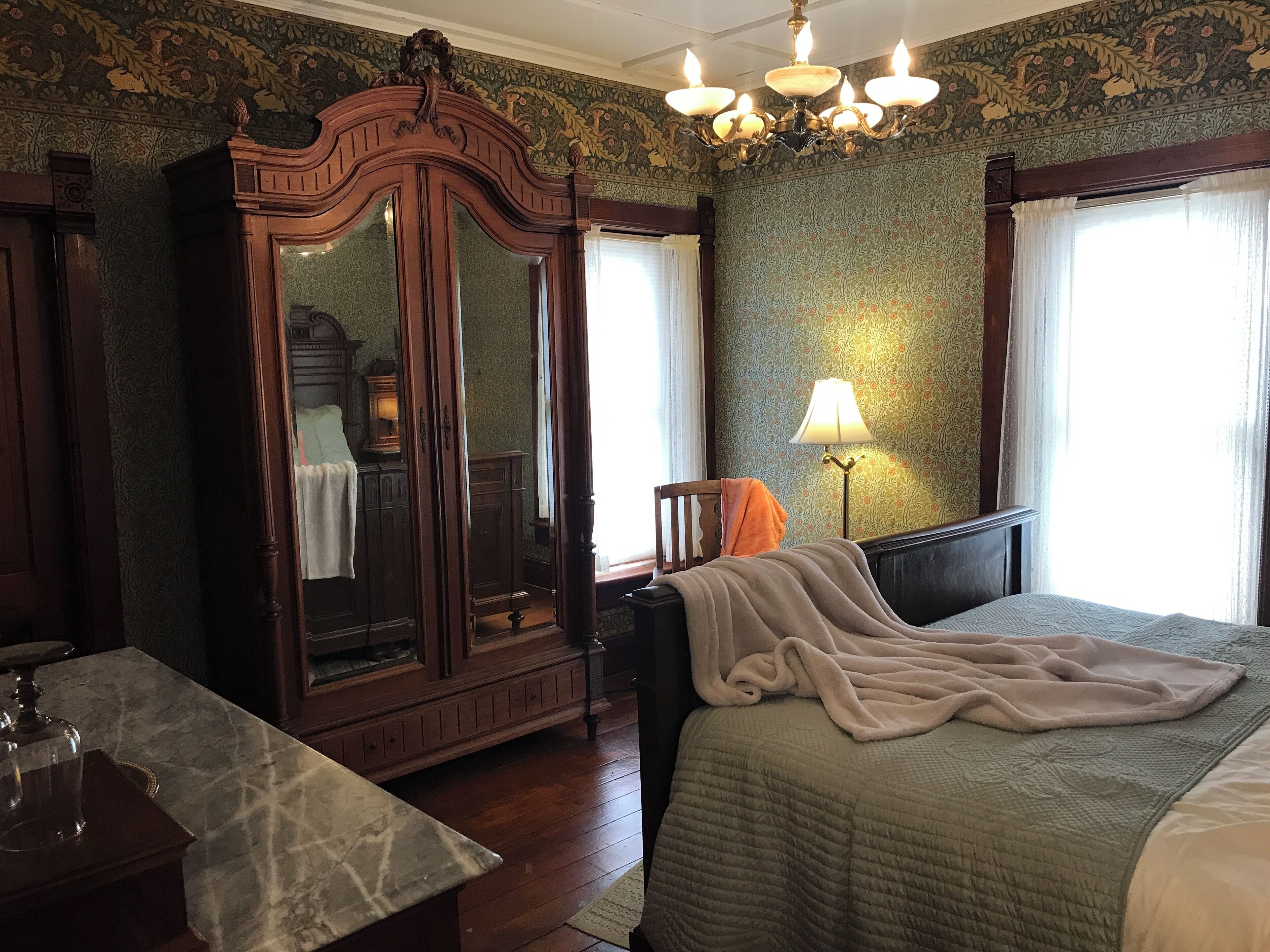 A bedroom with a large wooden wardrobe, a bed with a beige blanket, a nightstand with a lamp, and a window with curtains.
