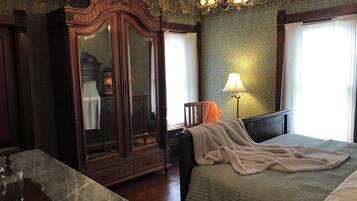 A bedroom with a large wooden wardrobe, a bed with a beige blanket, a nightstand with a lamp, and a window with curtains.