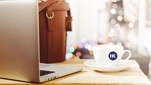 Un portatile su un tavolo di legno, con una tazza bianca recante la scritta 'HK' e una borsa in pelle marrone sullo sfondo.