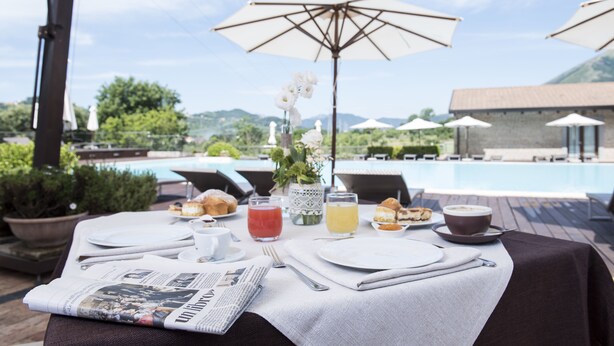 Mesa de desayuno al aire libre con periódico, bebidas, pasteles y vistas a una piscina y montañas.