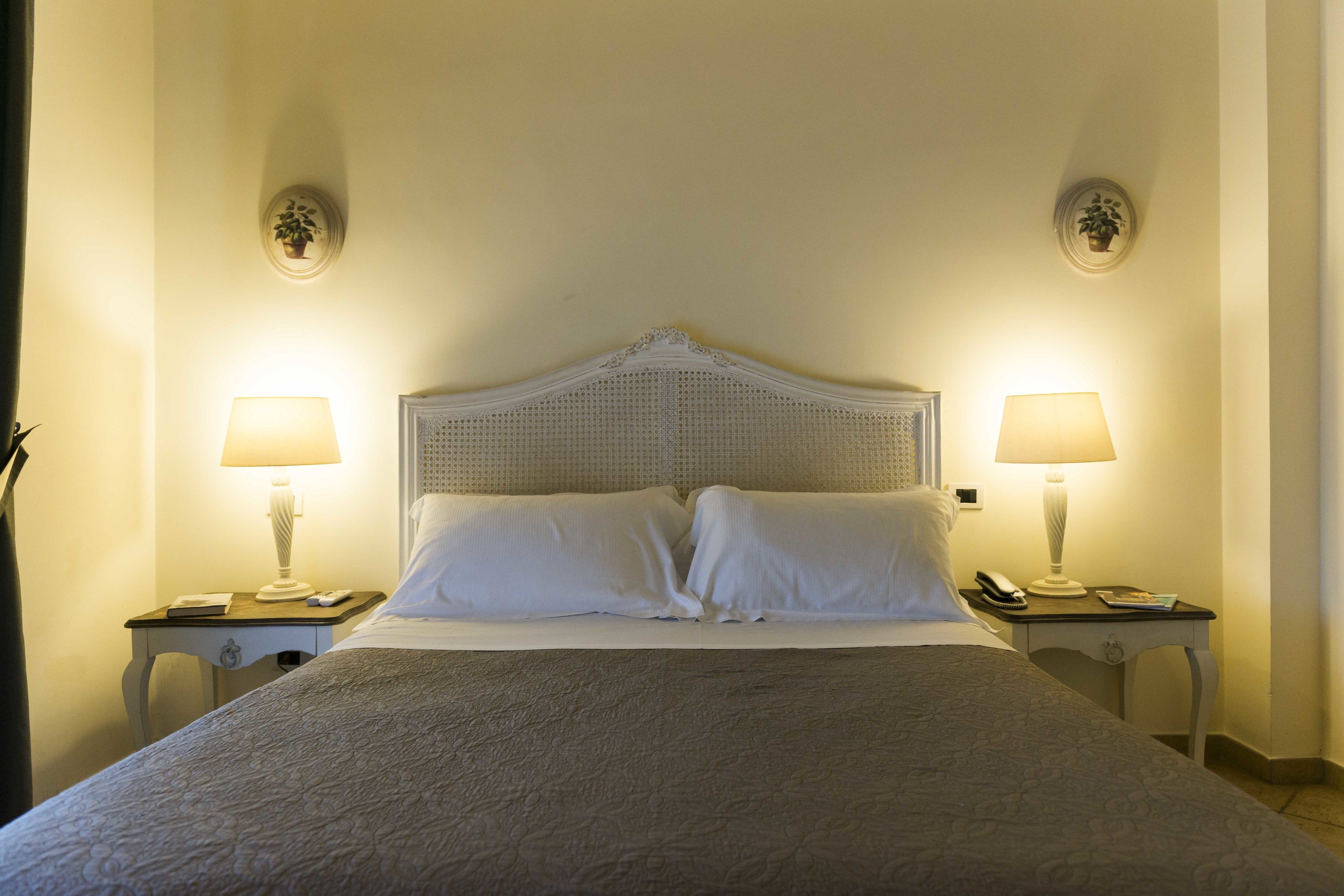Una cama bien hecha con sábanas blancas, dos mesitas de noche con lámparas, e iluminación empotrada en la pared.