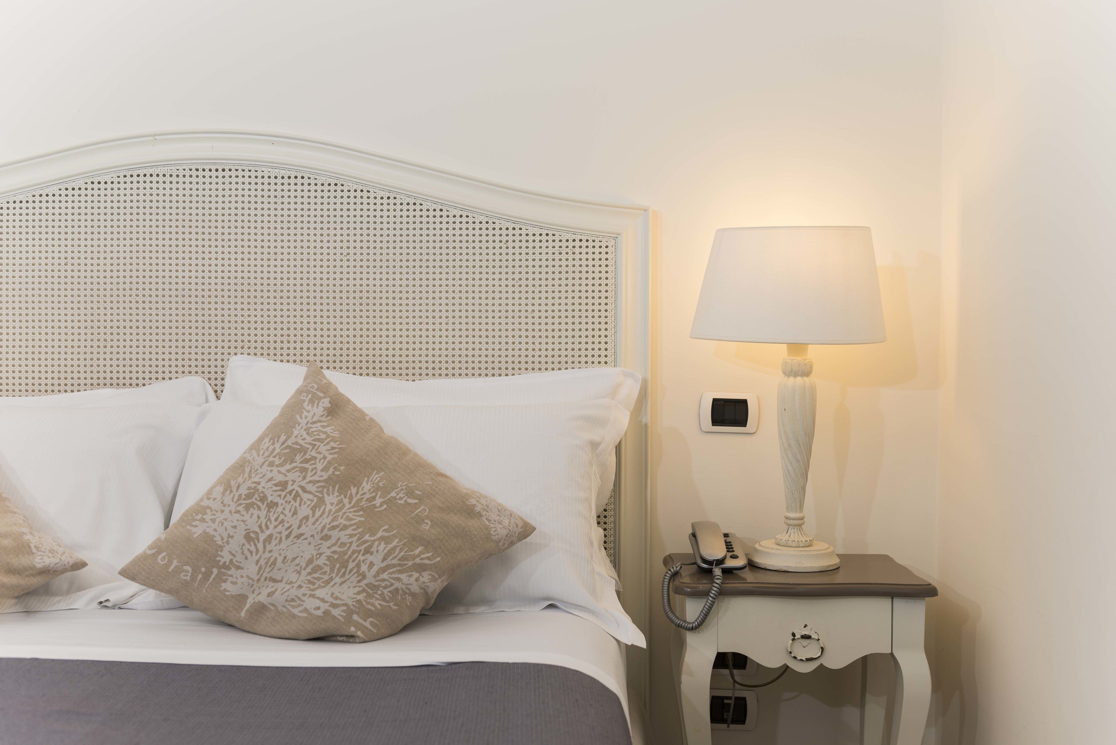 Un lit bien fait, avec des oreillers blancs et beiges, une tête de lit blanche et une table de chevet sur laquelle se trouvent une lampe et un téléphone.