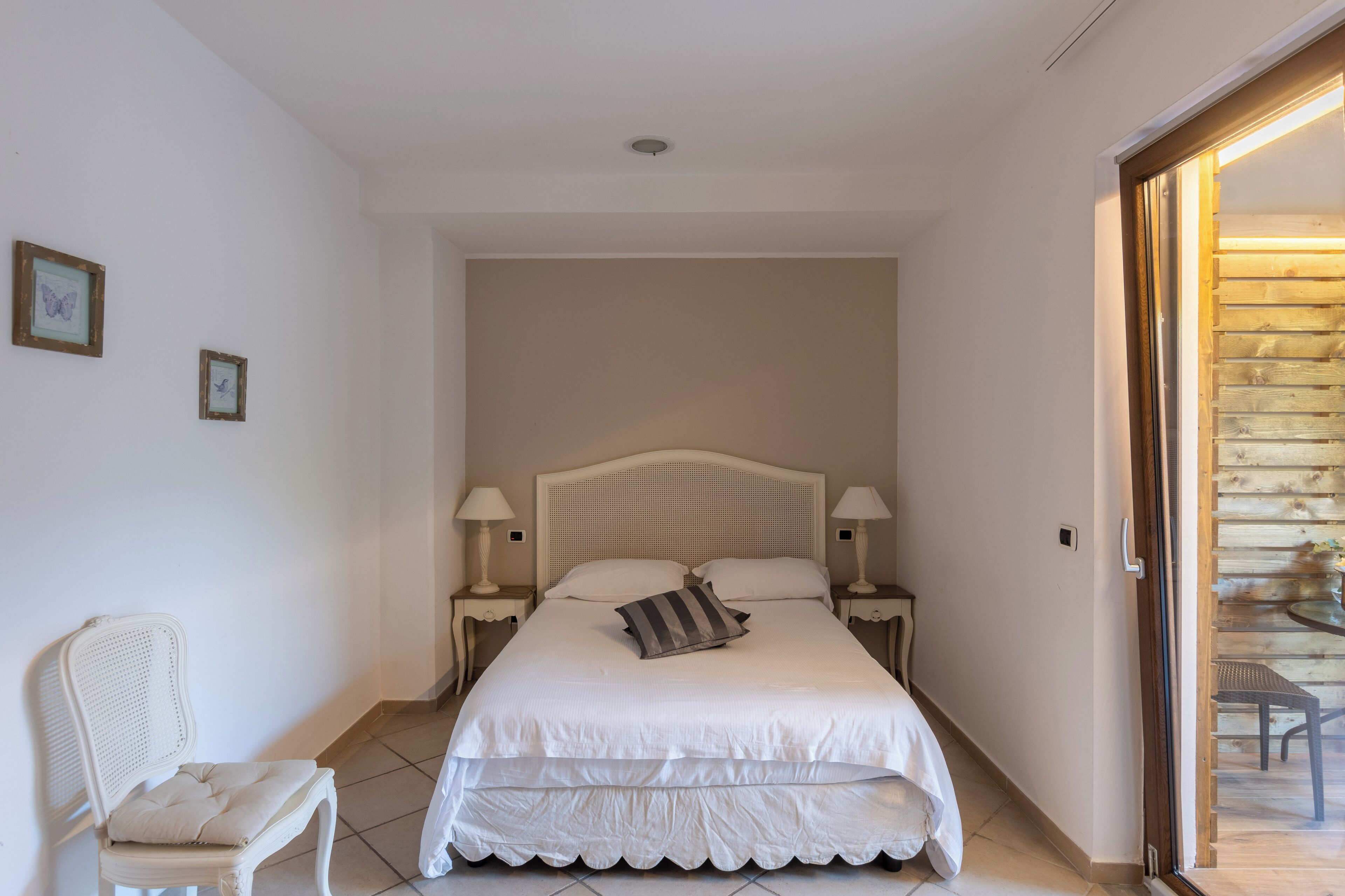 A bedroom with a large bed, two bedside tables with lamps, a chair, and a wooden door leading to an outdoor area.