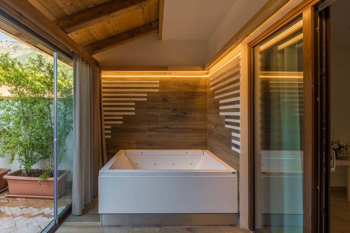A modern bathroom with a freestanding bathtub, wooden ceiling beams, and a view of greenery outside.
