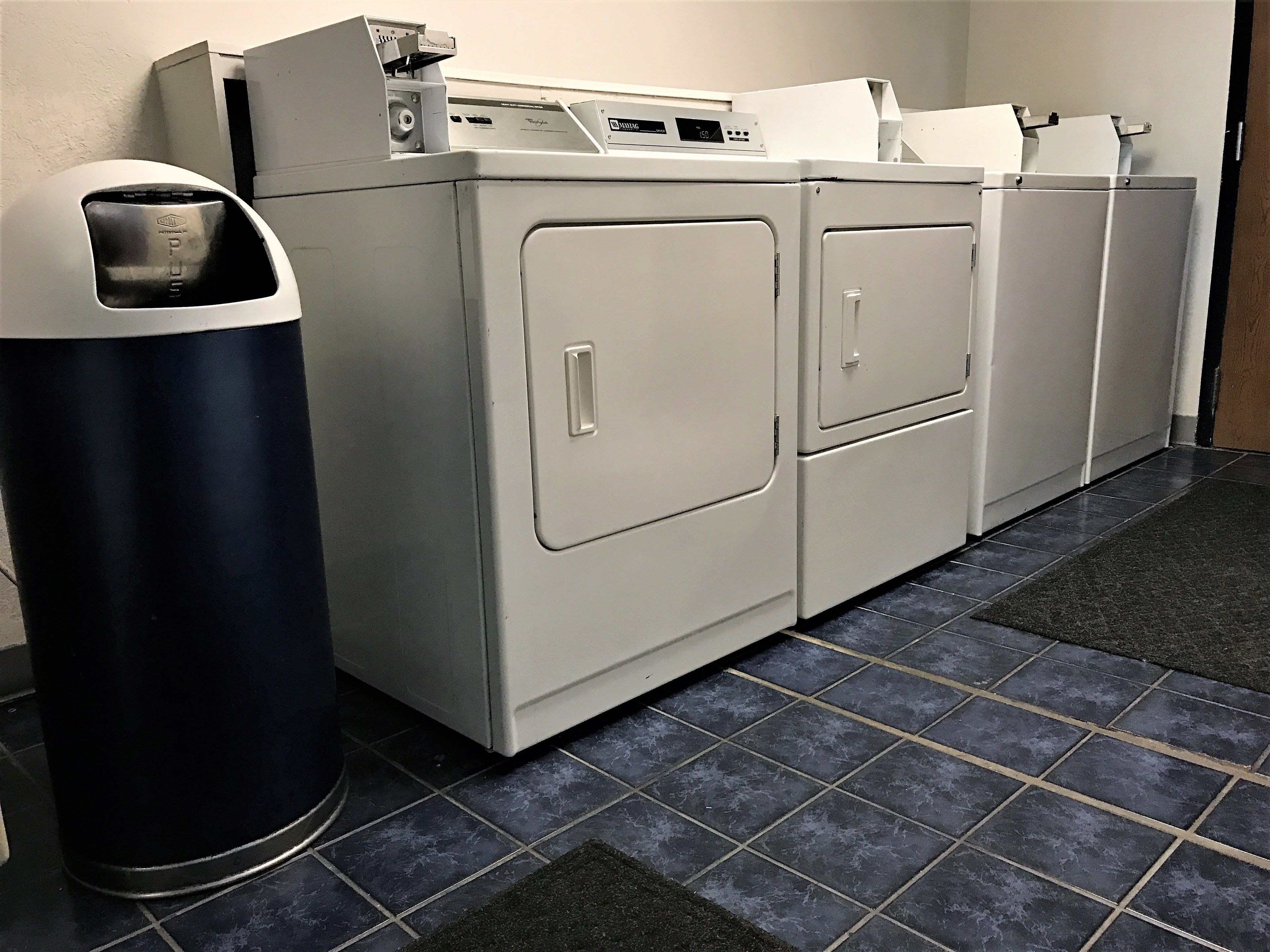 A laundry room with a front-loading washer and dryer, a trash can, and a tiled floor.
