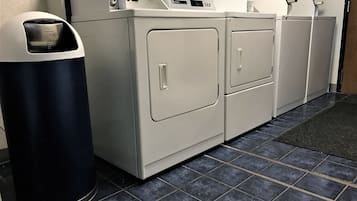 A laundry room with a front-loading washer and dryer, a trash can, and a tiled floor.