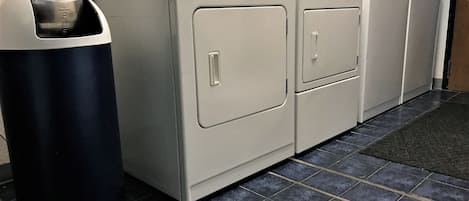 A laundry room with a front-loading washer and dryer, a trash can, and a tiled floor.
