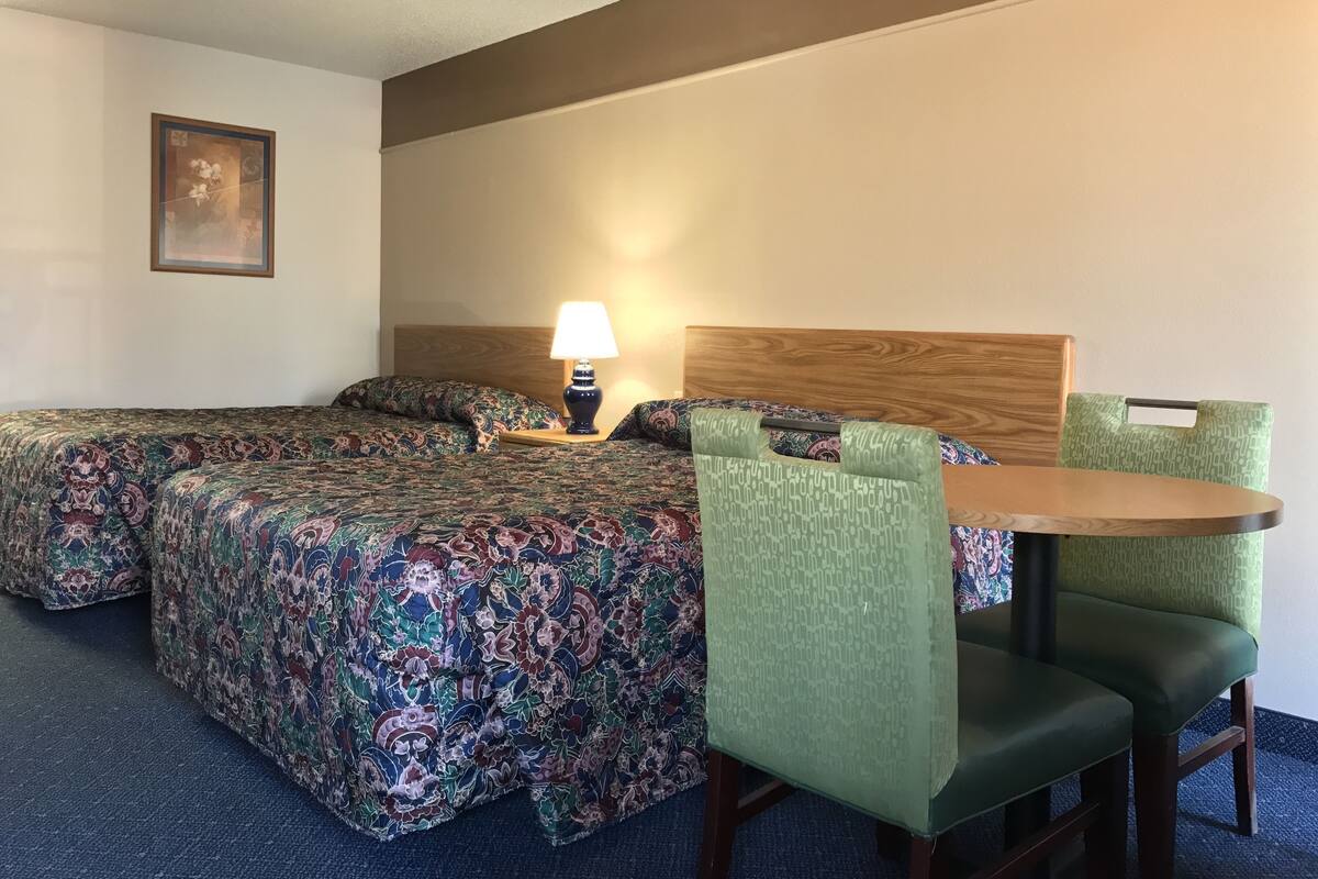 Een hotelkamer met twee bedden, een houten hoofdbord, een tafel met stoelen, een lamp en een lijst met een afbeelding aan de muur.