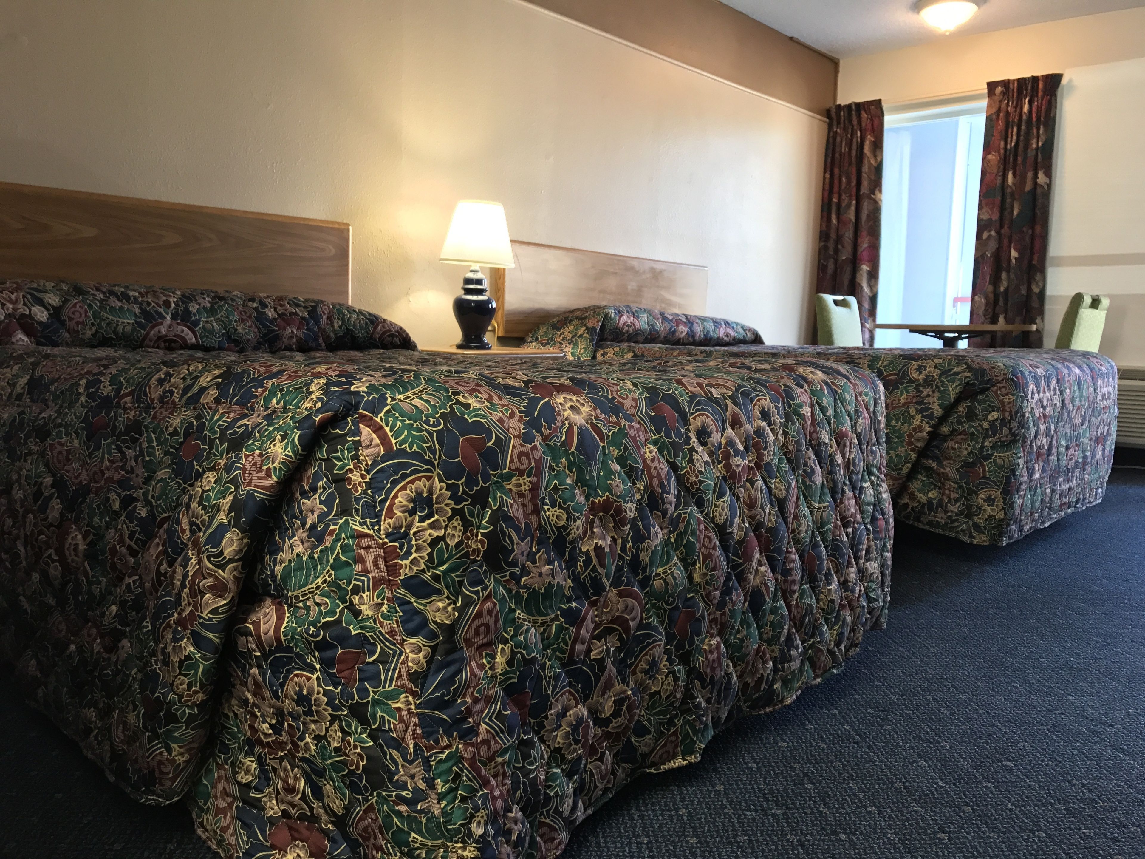 Ett hotellrum med två sängar, ett nattduksbord med en lampa, ett fönster med gardiner och en sänggavel i trä.