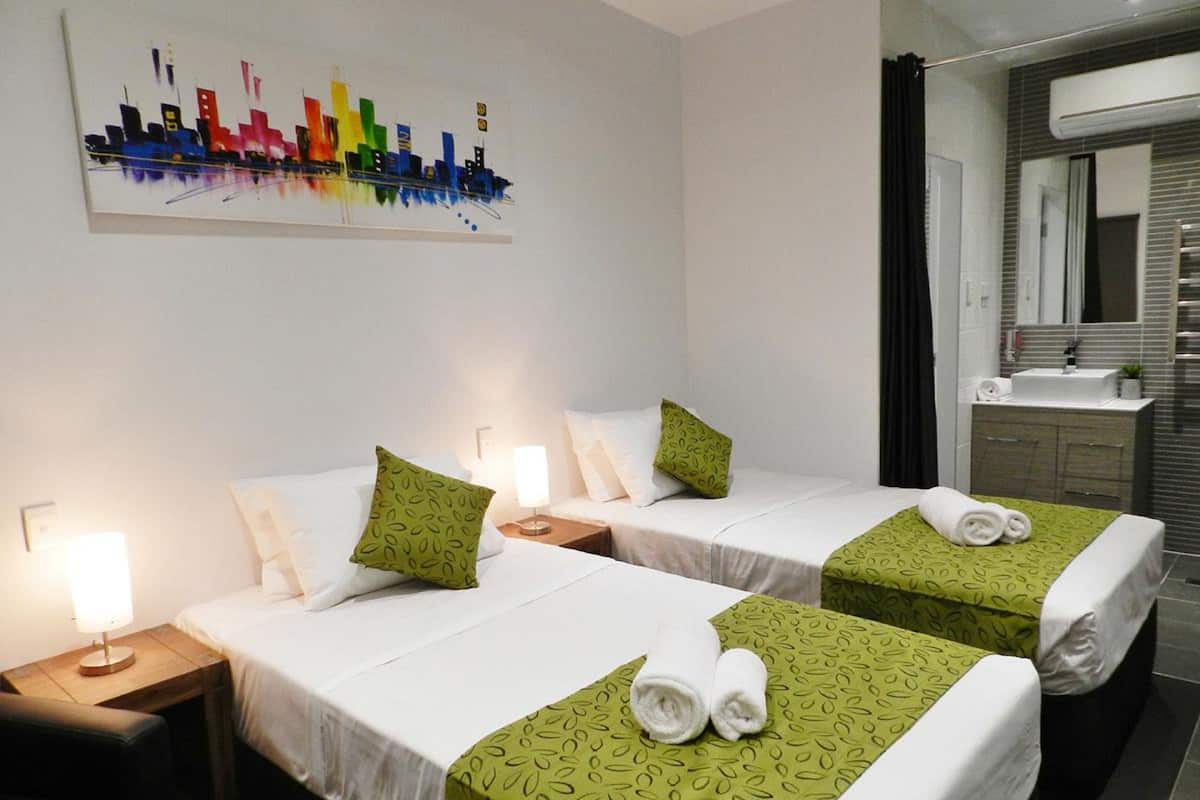A hotel room with two beds, each with a green patterned bedspread and white linens. There is a cityscape painting on the wall, a bathroom with a mirror and sink, and a nightstand with a lamp.