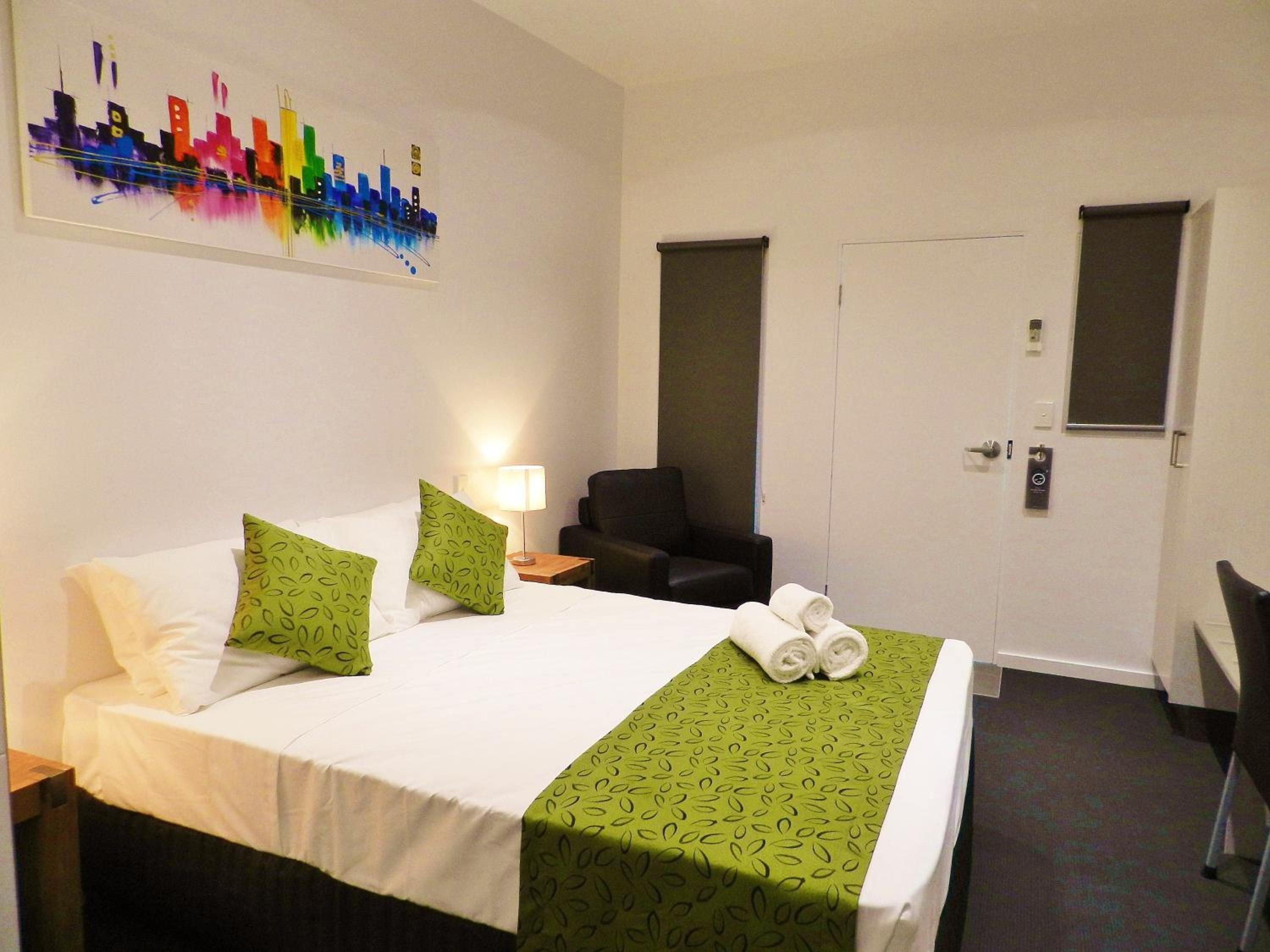 Hotelkamer met een bed, groene kussens met een patroon, een groen dekbed, een stoel, een bureau, een lamp en een deur met een raam.
