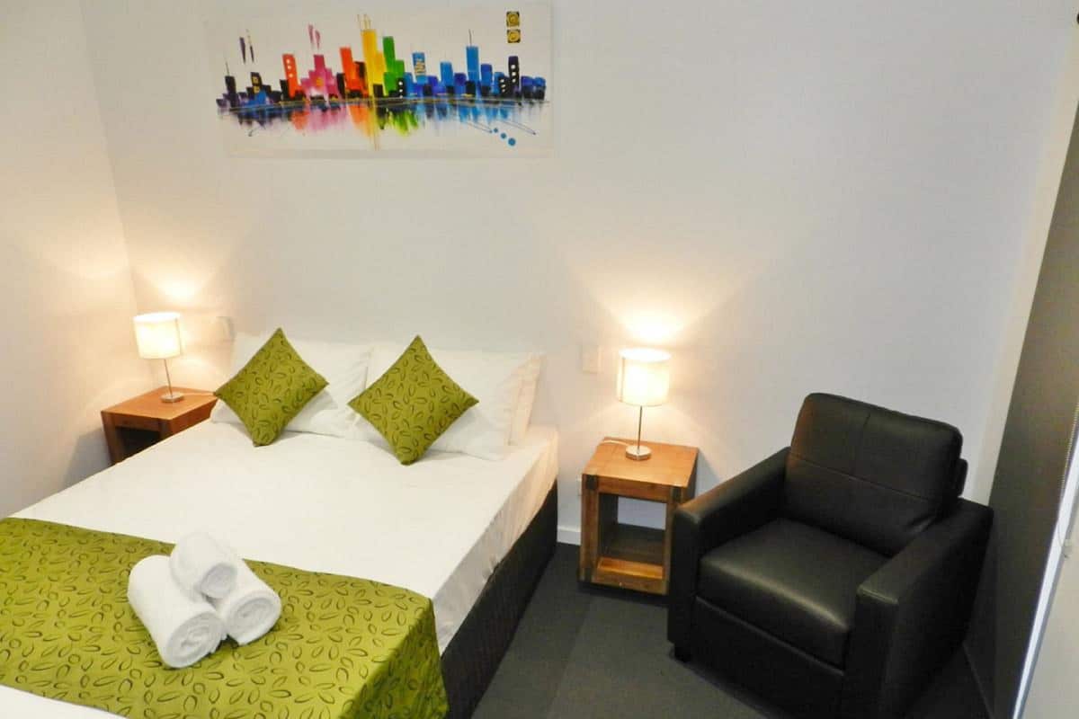 A hotel room with a bed, two nightstands, a chair, and a cityscape artwork on the wall.