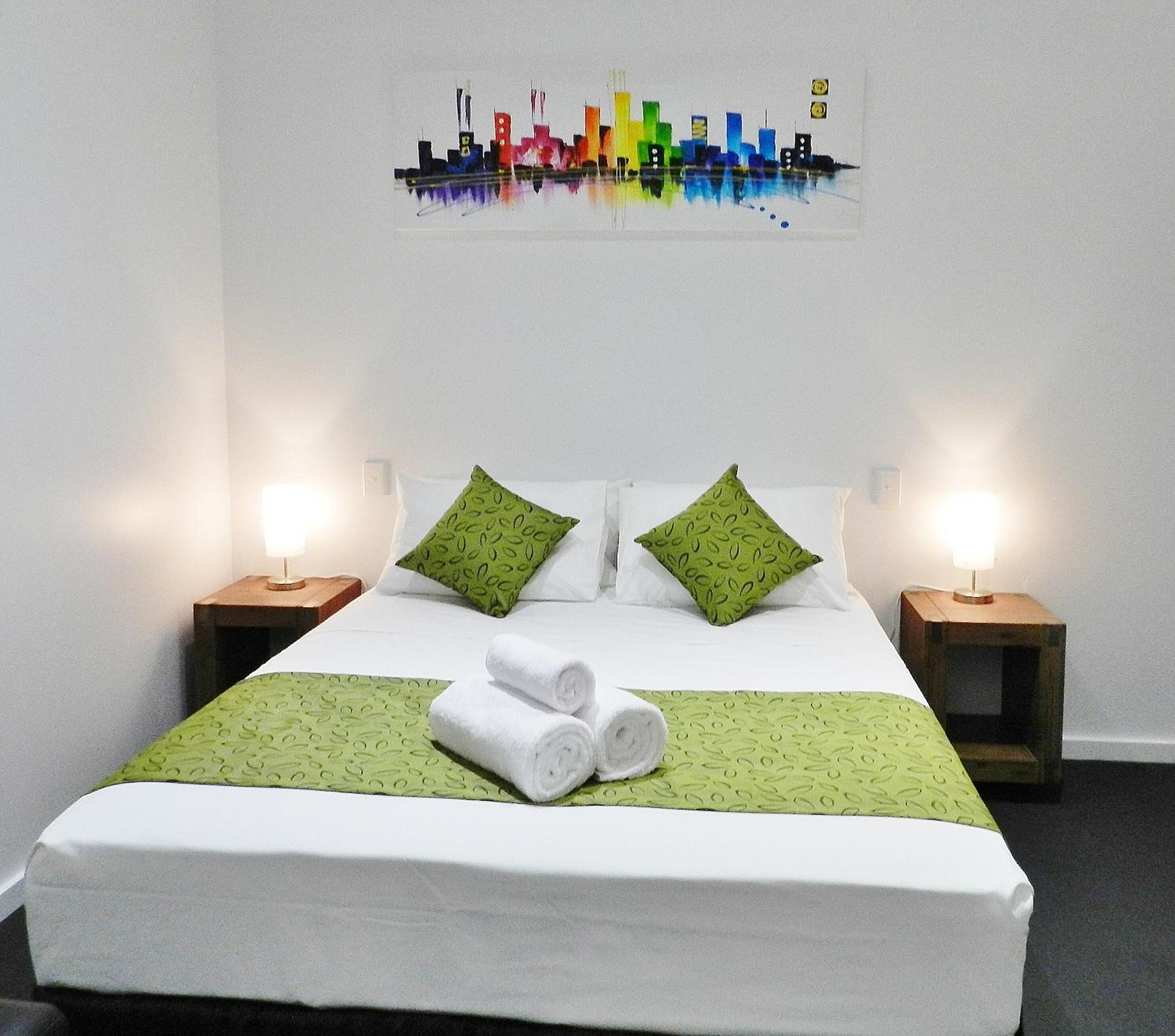 A neatly made bed with green pillows and a green bedspread, two bedside tables with lamps, and a colorful cityscape painting on the wall.