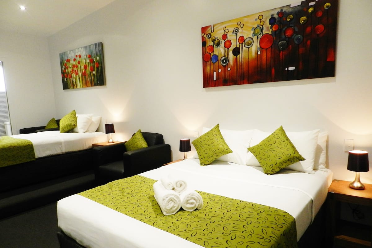 A hotel room with two beds, green patterned bedspreads, white pillows, and green throw pillows. There are two paintings on the walls, a lamp on each bedside table, and a folded towel on the bed.