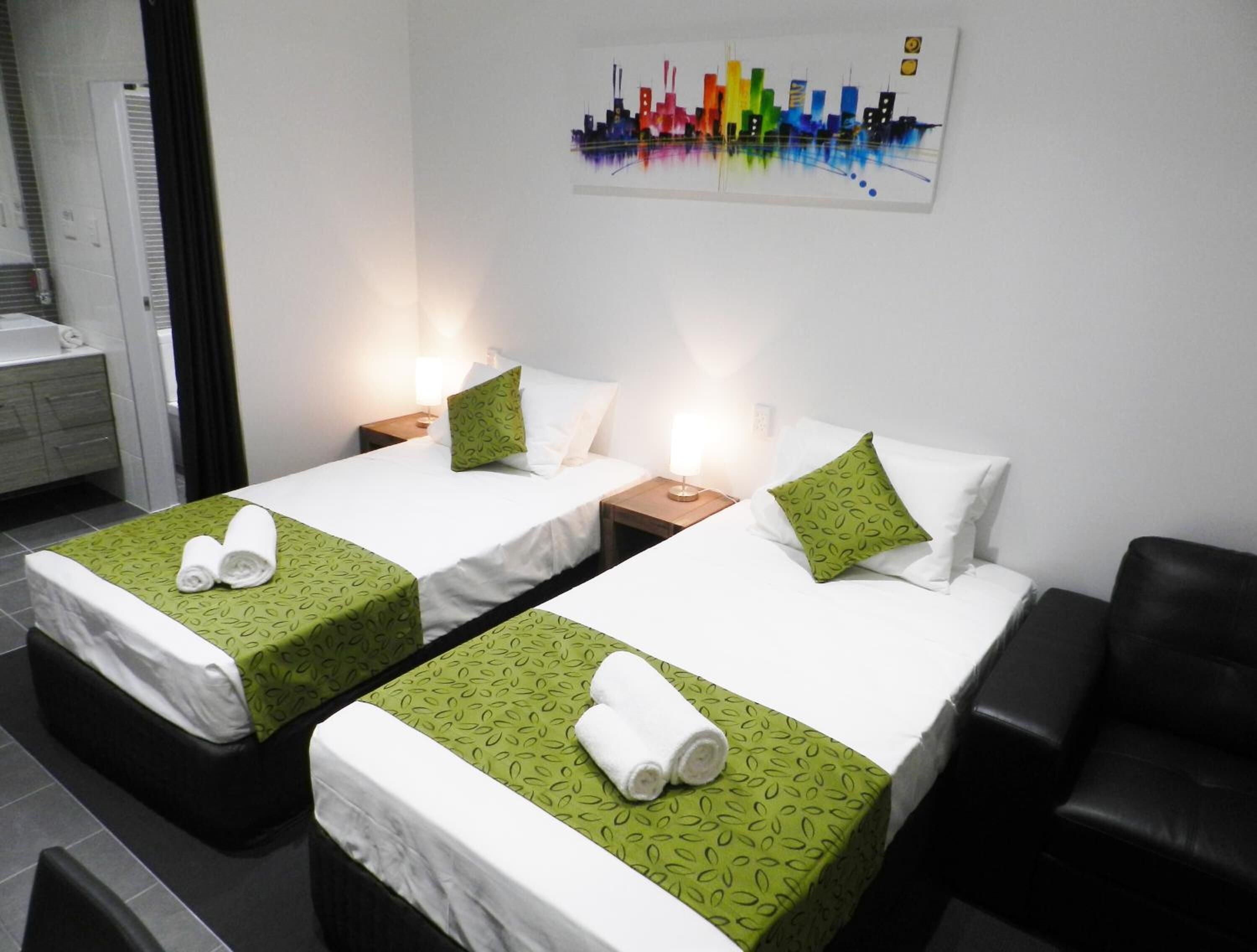 Ein Hotelzimmer mit zwei Betten, jeweils mit einem grün gemusterten Bettbezug und weißen Bettwäsche, einem schwarzen Ledersessel und einem Stadtbild an der Wand.