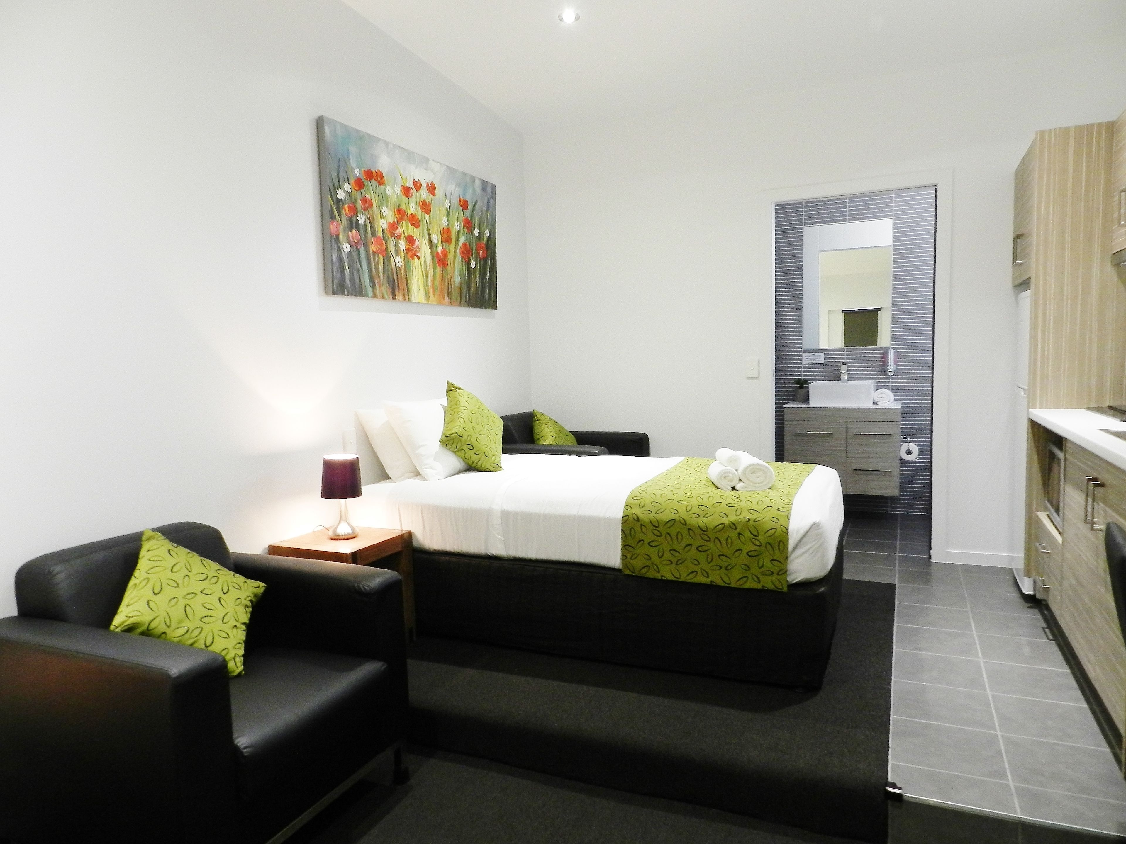 A modern hotel room with a bed, a bedside table with a lamp, a green patterned pillow, a painting on the wall, and a bathroom visible through an open door.