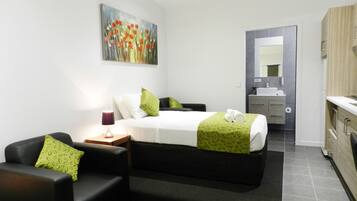 A modern hotel room with a bed, a bedside table with a lamp, a green patterned pillow, a painting on the wall, and a bathroom visible through an open door.