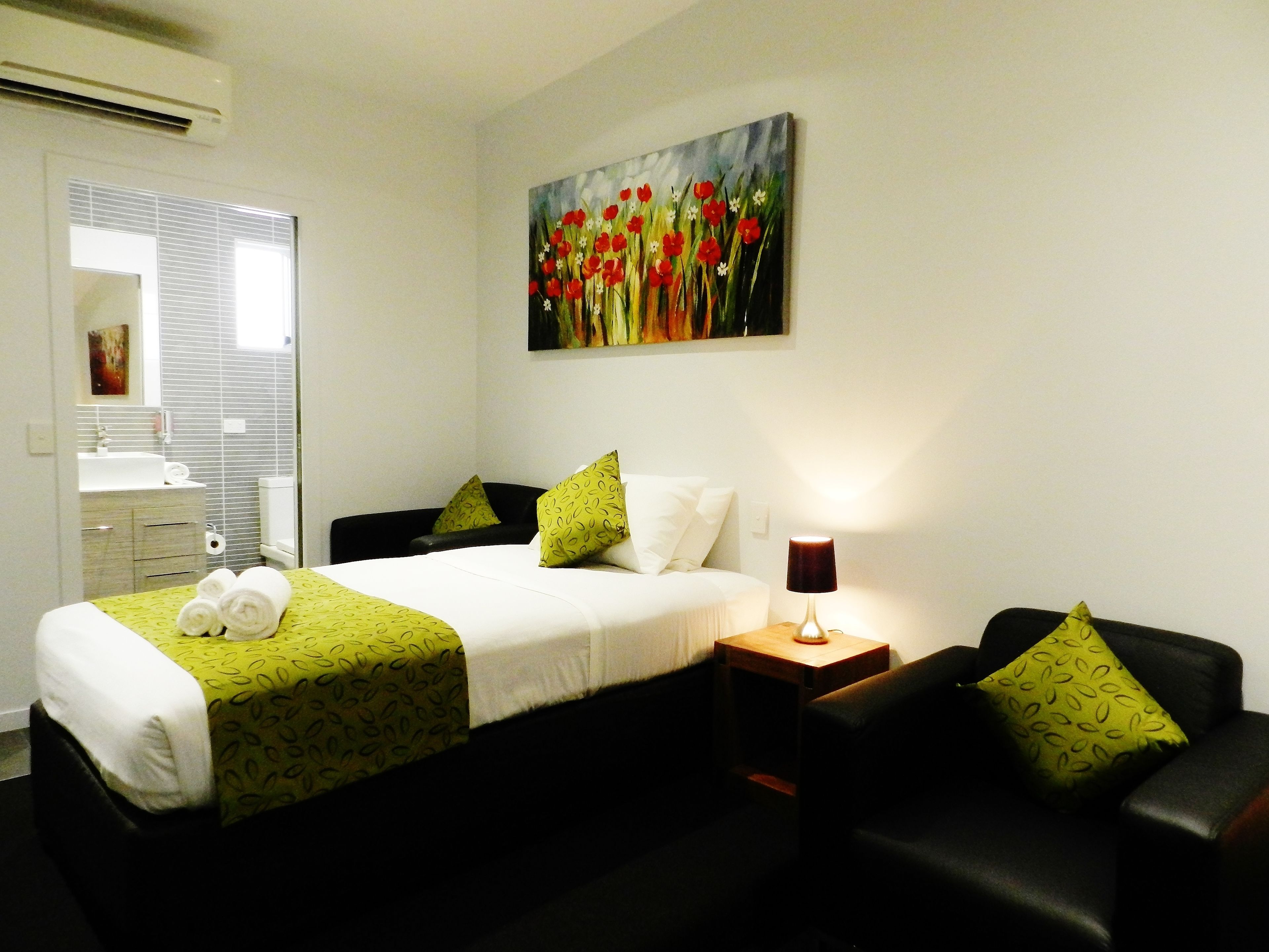 A hotel room with a bed, a bedside table with a lamp, a black leather armchair, a painting on the wall, and a bathroom visible through an open door.