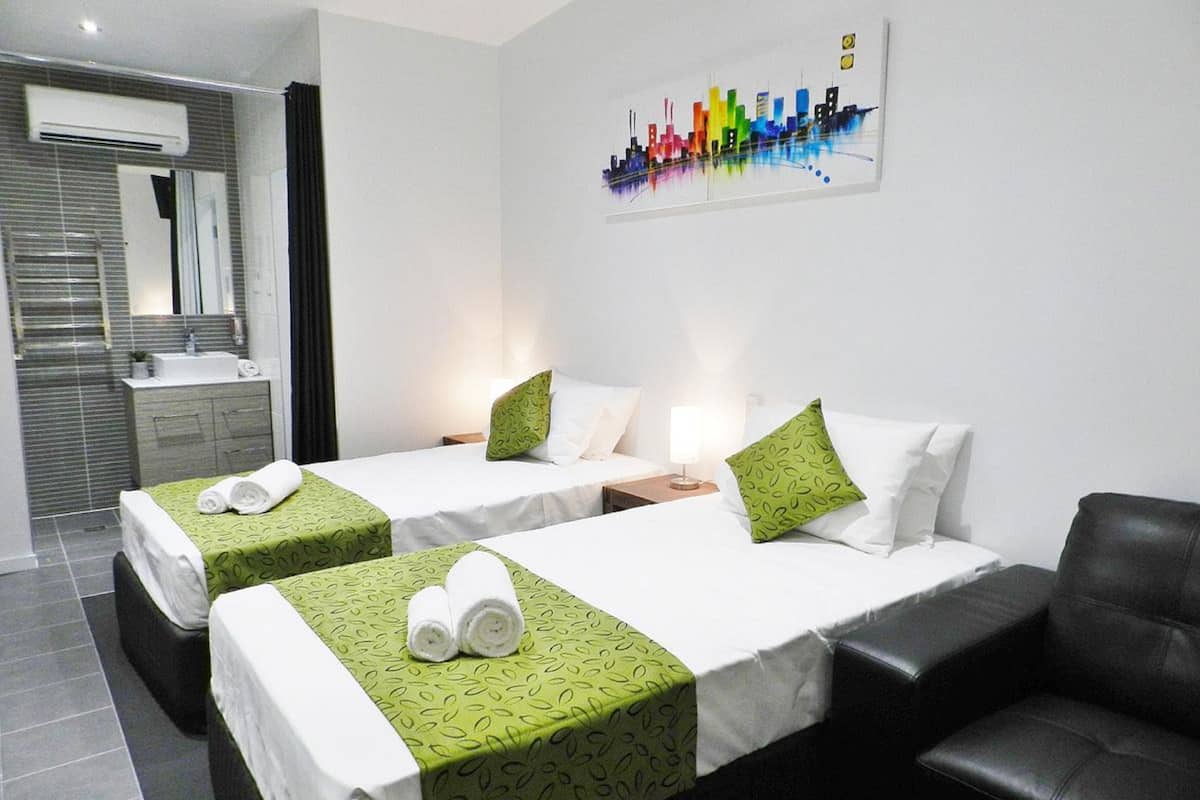 A hotel room with two beds, a sofa, a bathroom with a mirror, and a cityscape painting on the wall.