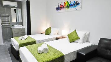 A hotel room with two beds, a sofa, a bathroom with a mirror, and a cityscape painting on the wall.