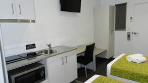 A compact hotel room with a kitchenette, a bed with green patterned bedding, a desk, and a TV mounted on the wall. - Copper City Motel (Pioneer)