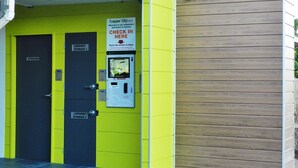 A hotel entrance with a bright yellow wall, a 'Check in Here' sign, and a self-check-in kiosk. - Copper City Motel (Pioneer)