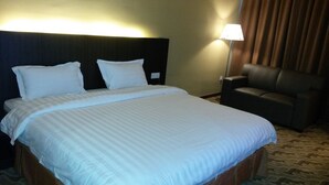 A hotel room with a large bed, two pillows, a nightstand, a lamp, and a leather armchair. - Eminent Hotel (Kota Kinabalu)