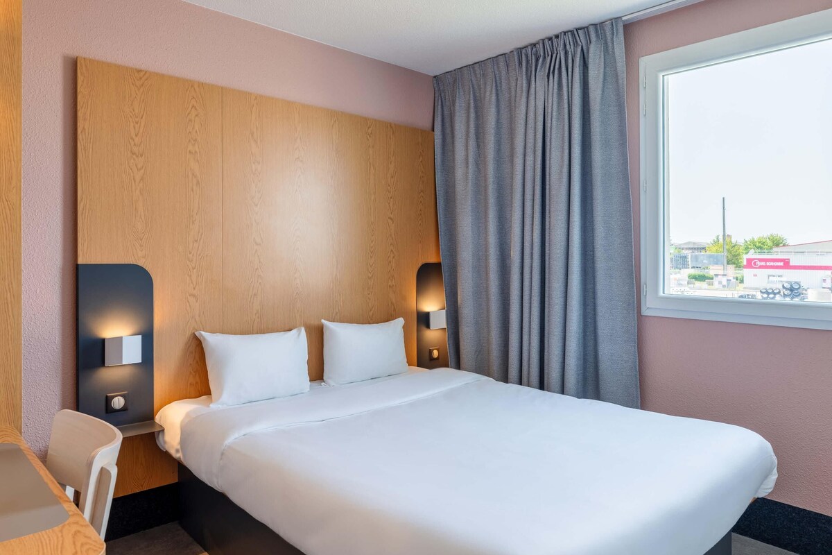 A hotel room with a large bed, two bedside lamps, a desk, and a window with curtains.