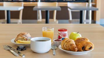 A breakfast setting with croissants, an apple, a glass of orange juice, a bowl of coffee, and condiments on a table.