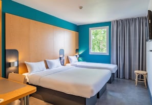 Ein Hotelzimmer mit zwei Betten, einem kleinen Tisch, einem Stuhl, einem Fenster mit Vorhängen und einem Fernseher.