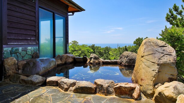 A stone-tiled hot tub surrounded by rocks, a wooden building with glass doors, and a view of the ocean. - Grampus Resort Shirahama (Shirahama)