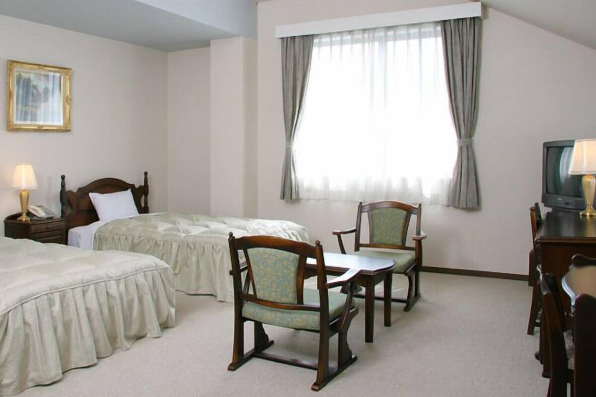 Quarto de hotel com duas camas de solteiro, uma mesa com televisão, uma cadeira e uma janela com cortinas.