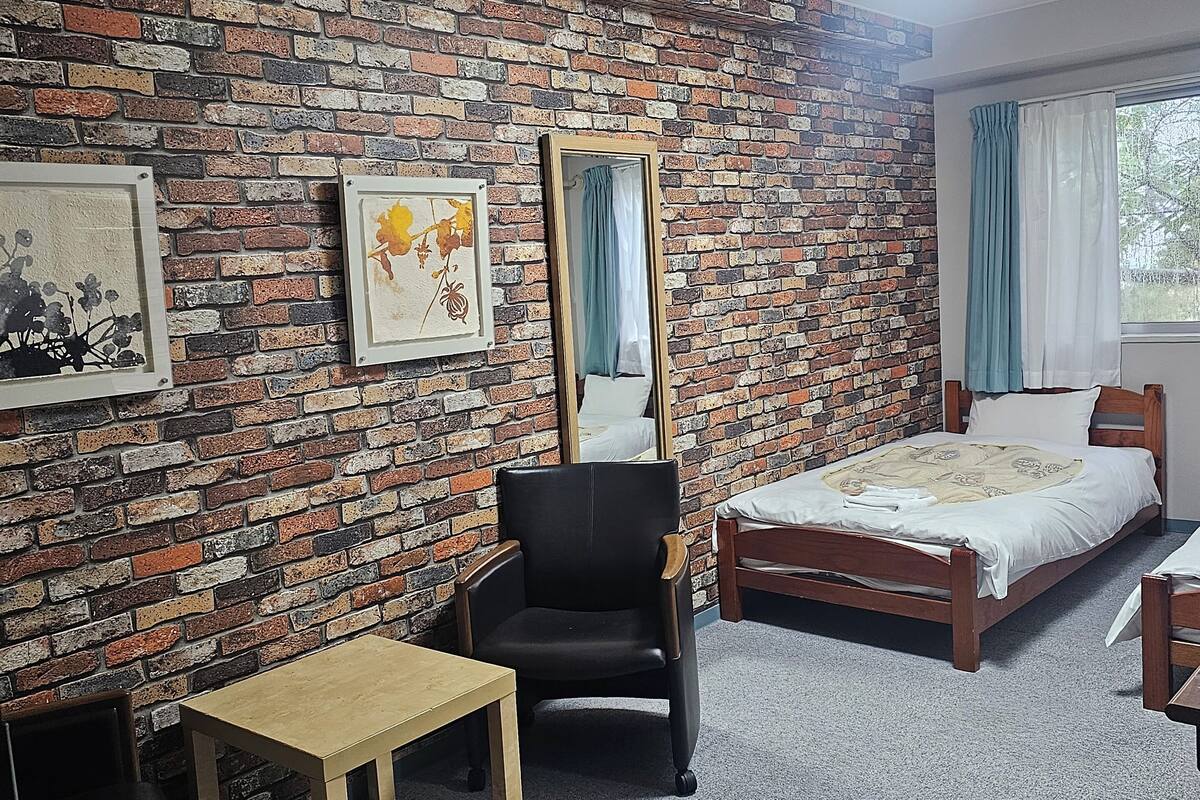 Quarto com parede de tijolos, uma cama, uma cadeira, uma mesinha e janela com cortinas.