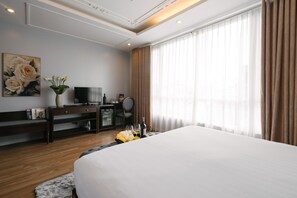 A hotel room with a large bed, a TV, a desk with a computer, a vase with flowers, and a window with curtains. - La Palm Hotel (Hanoi)