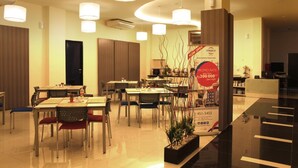A modern hotel dining area with multiple tables and chairs, a central promotional display, and a reception desk. - Cordela Hotel Medan (Medan)