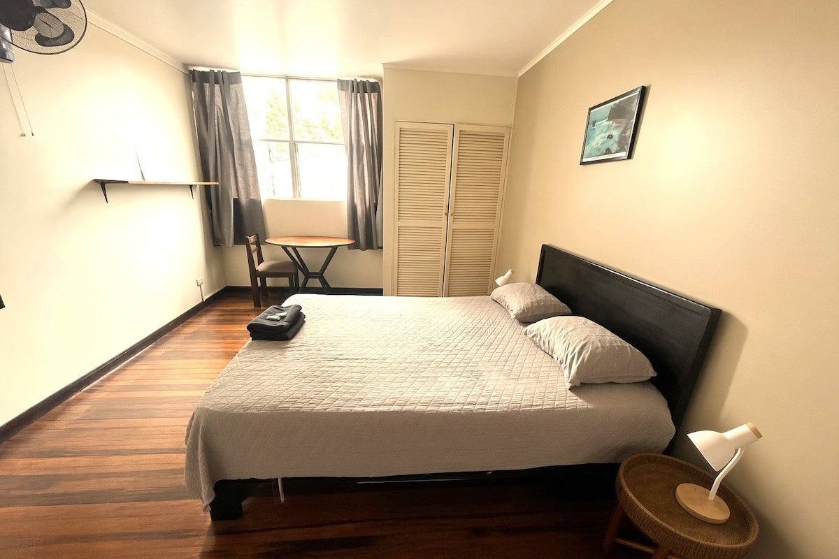 Un dormitorio con cama, una silla, una mesa, una lámpara y una ventana con cortinas.