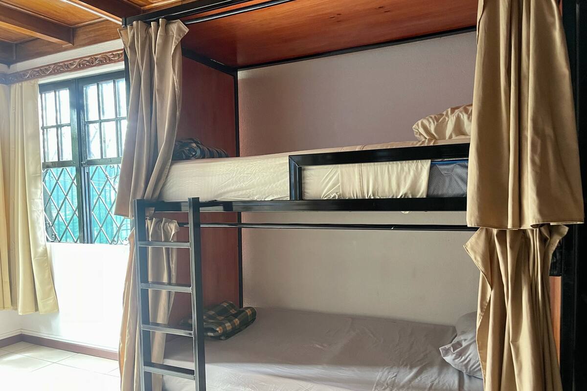Une chambre avec un lit superposé, une échelle, une fenêtre avec des barreaux en métal et des rideaux.
