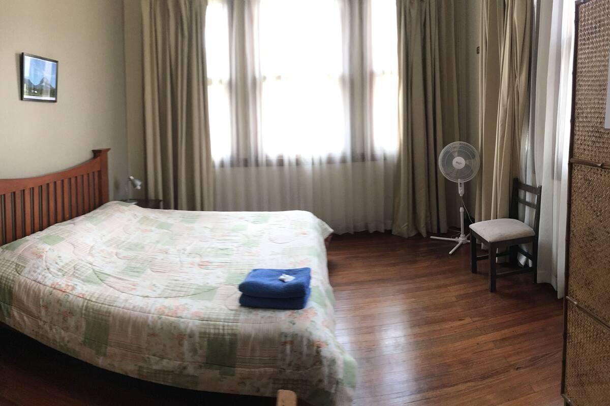 Une chambre à coucher bien rangée, avec un lit en bois, une lampe de chevet, une chaise et un ventilateur sur pied.