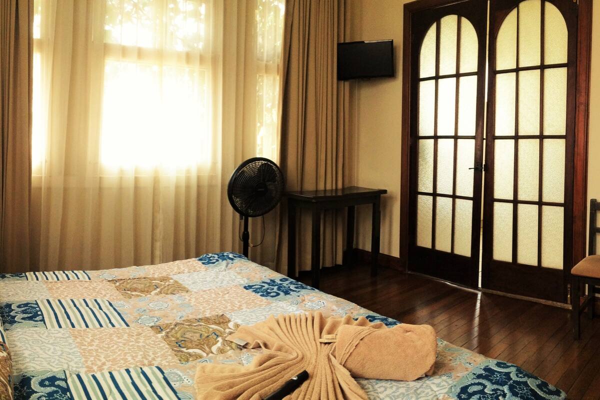 Une chambre d’hôtel avec un grand lit, un ventilateur, une chaise et une porte en bois dotée de panneaux de verre.