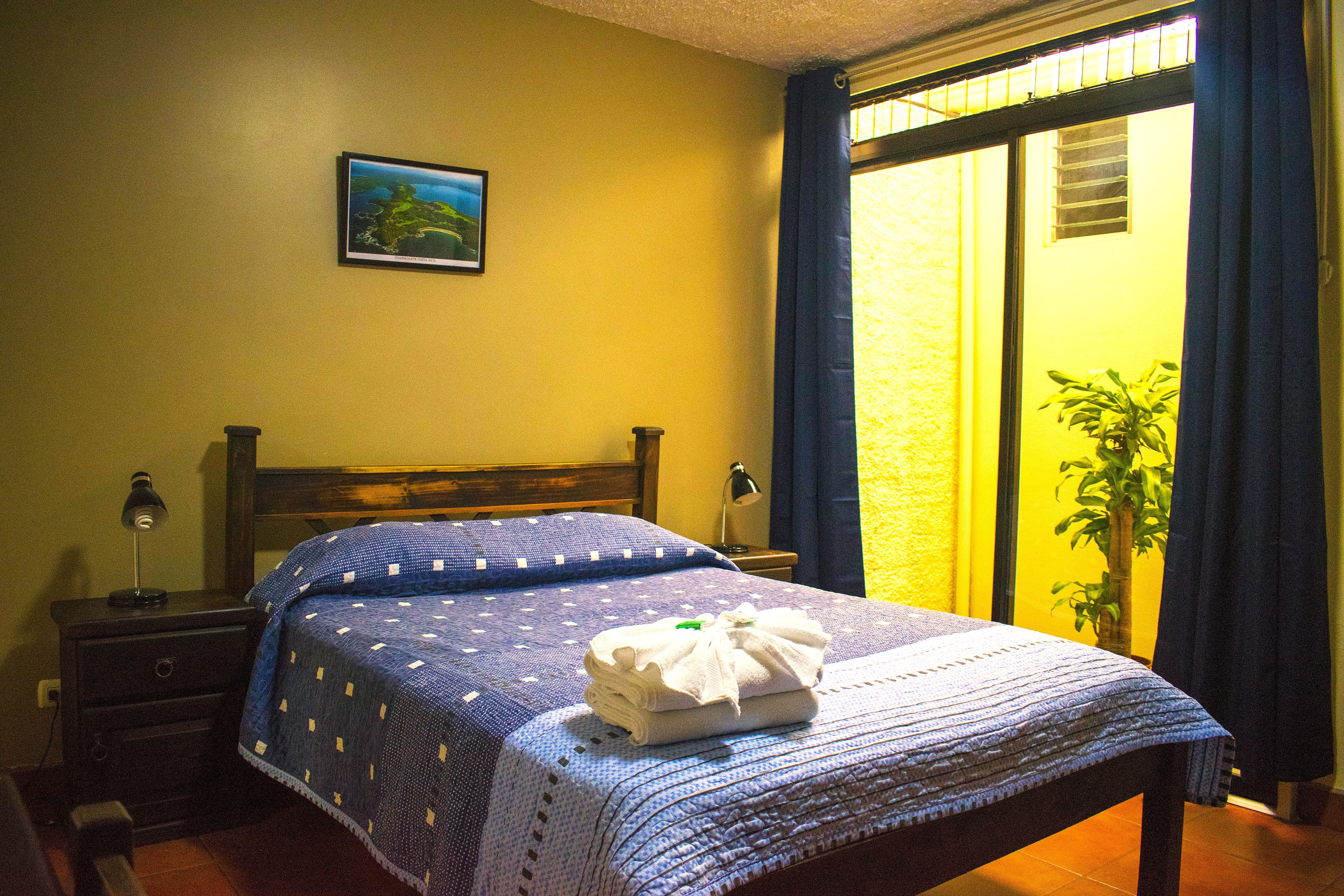 Cama bem arrumada com colcha azul, abajur e mesa pequena com luminária.