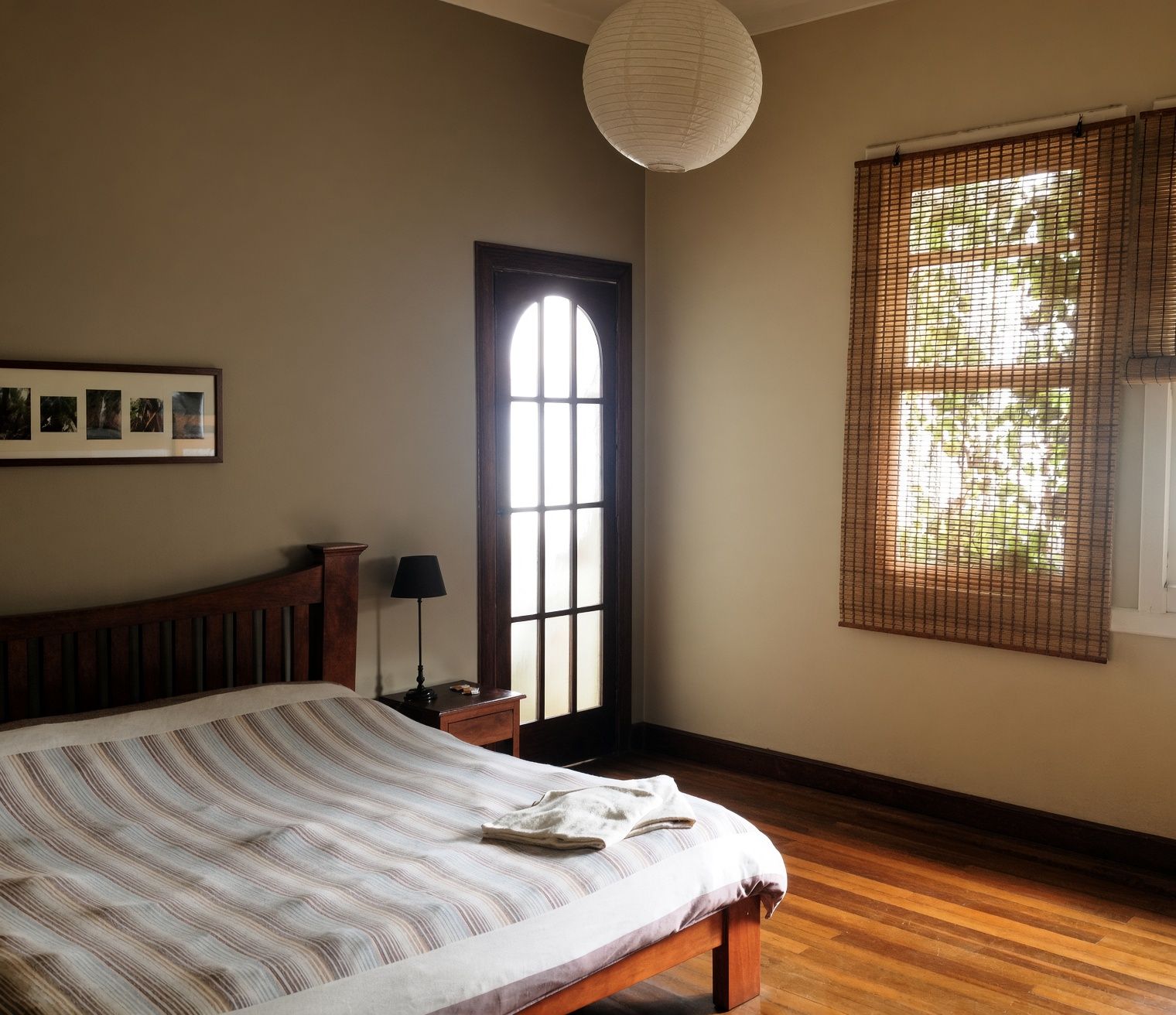 A bedroom with a large bed, a wooden headboard, a nightstand with a lamp, a framed picture on the wall, a window with a view, and a hanging light fixture.