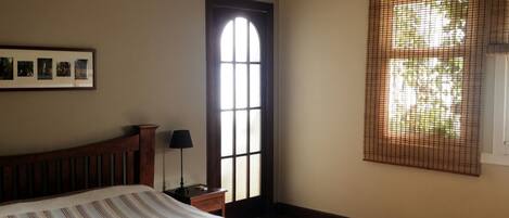 A bedroom with a large bed, a wooden headboard, a nightstand with a lamp, a framed picture on the wall, a window with a view, and a hanging light fixture.