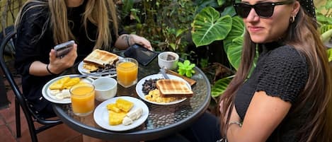 Dos personas sentadas en una mesa al aire libre con varios alimentos para el desayuno, rodeadas de follaje.