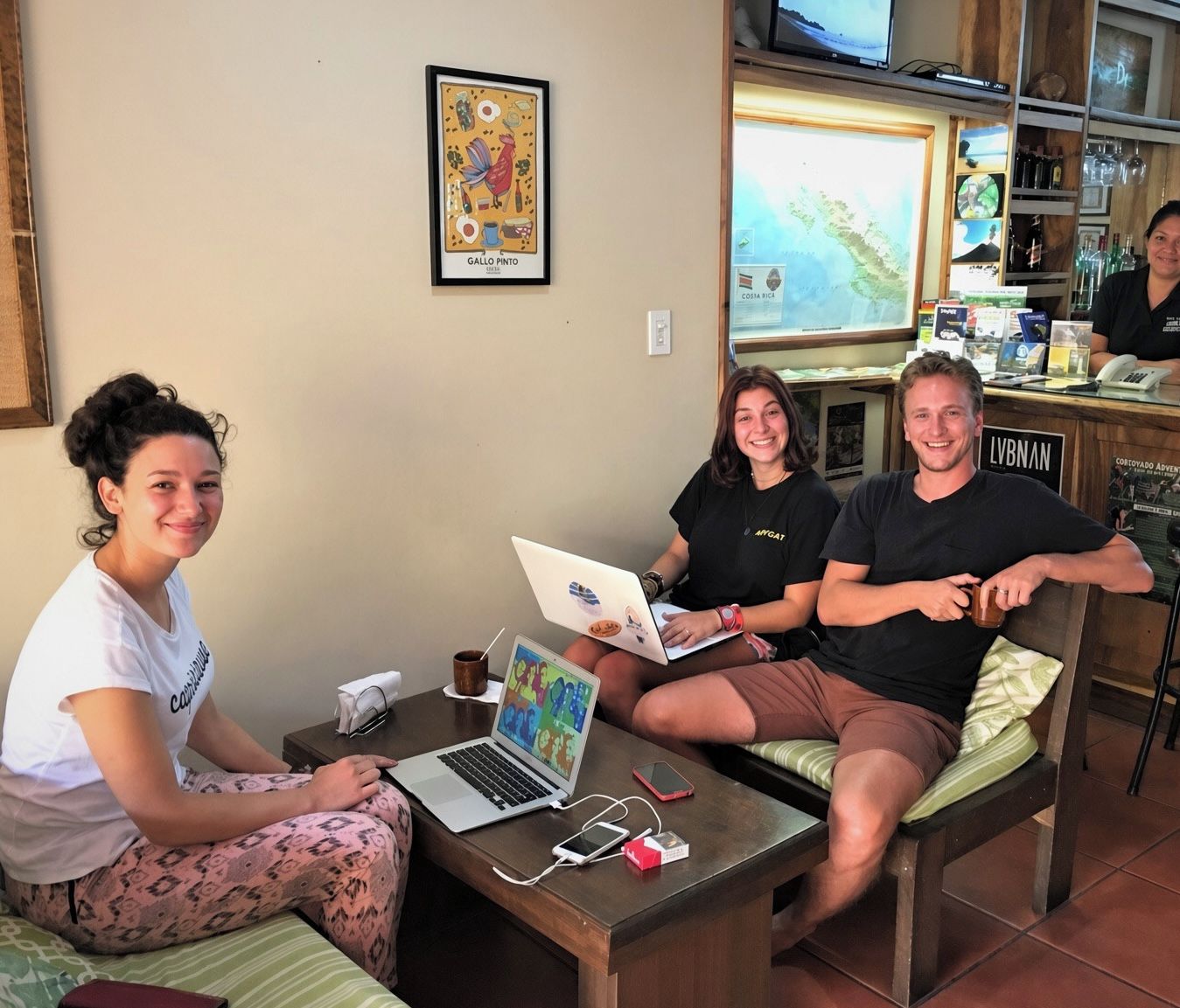 A group of people in a room with a laptop, a smartphone, and a tablet on a table. There is a framed map on the wall and a television on a shelf.