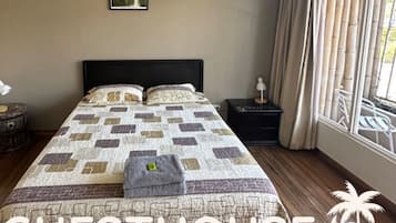 Un dormitorio con una cama grande, dos mesitas de noche, un ventanal con cortinas y dos cuadros en la pared.
