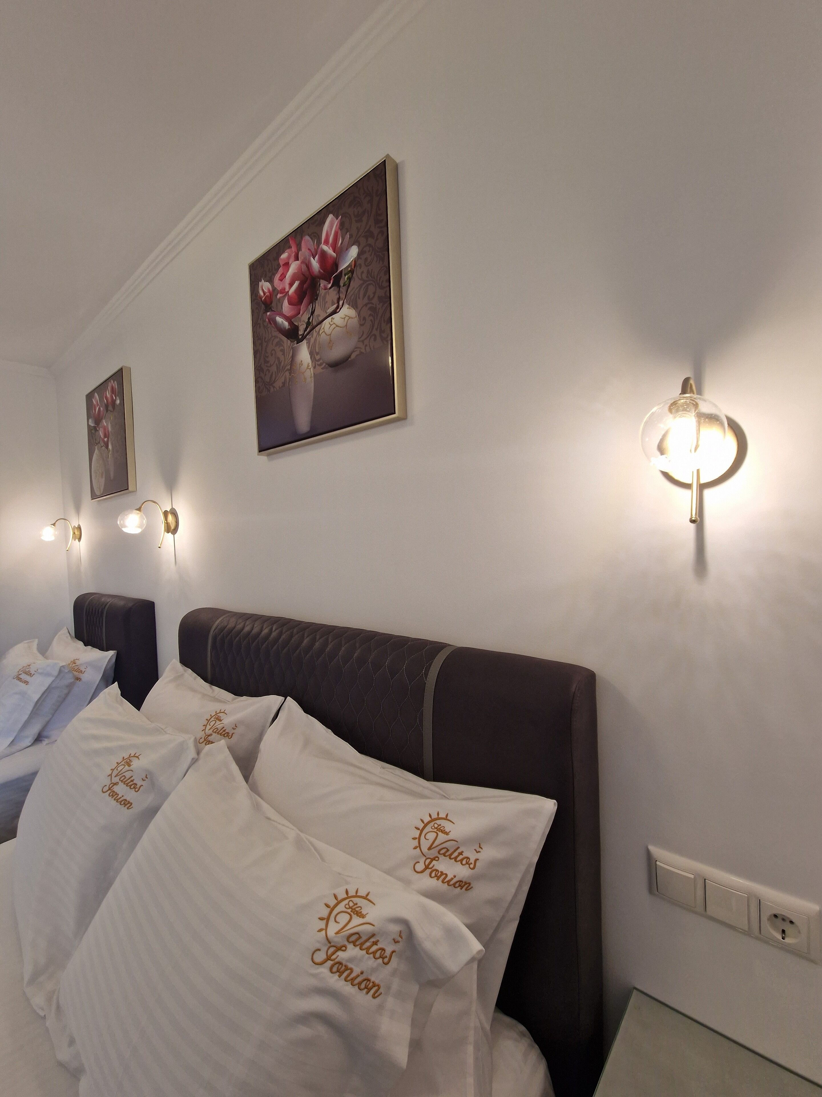A hotel room with a bed, white pillows with embroidered text, a wall-mounted light fixture, and framed floral artwork.